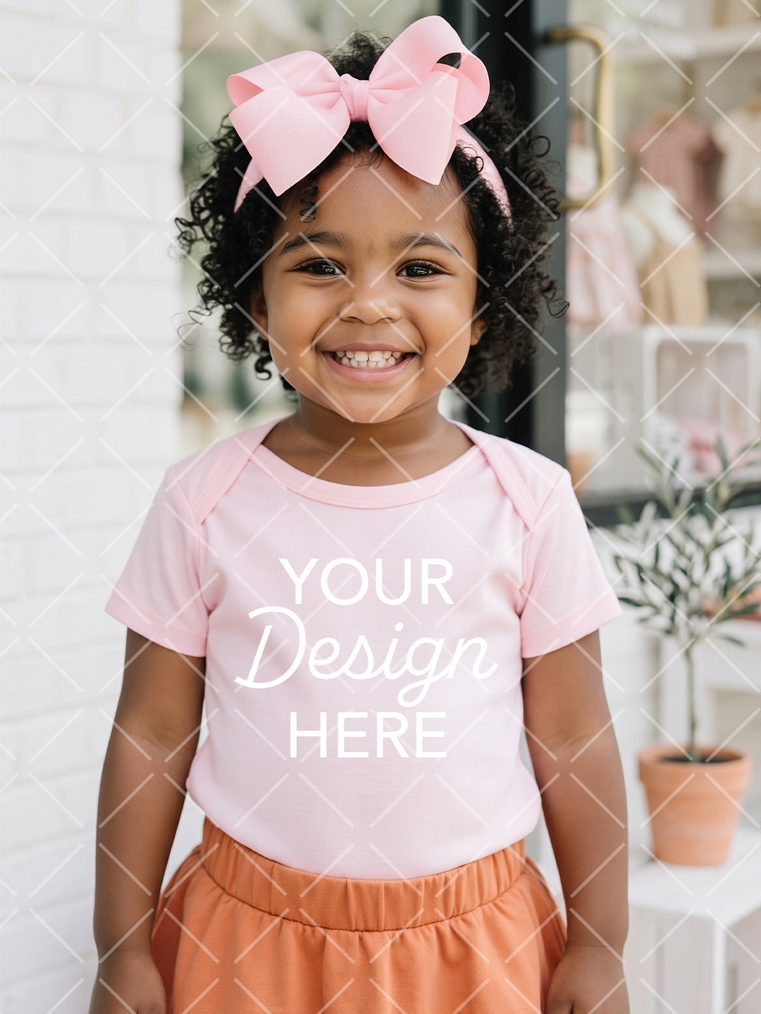 Smiling little girl wearing a light pink T-shirt, oversized pink hair bow, and orange shorts in a softly styled boutique setting.